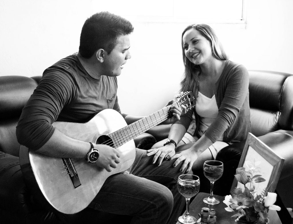 Imagem de um casal de homem e mulher. Eles estão em um sala, sentados cada um do lado do outro. Ele canta para ela e toca uma música em seu violão.