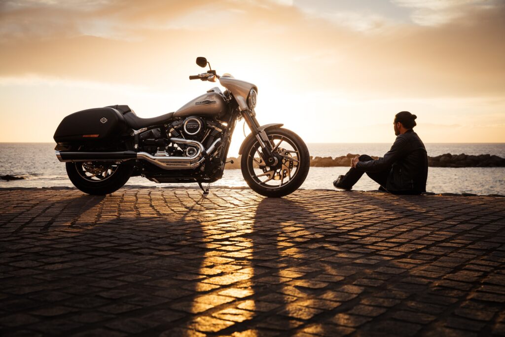 Imagem de um homem sentado em uma calçada de paralelepípedo. Ao lado dele uma linda moto. Ele está olhando para o infinito azul do mar.
