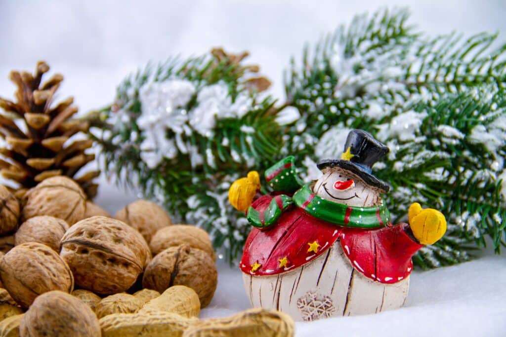 Imagem de nozes sobre uma mesa branca decorada com enfeites natalinos como pinha, boneco de neve e galho de um pinheiro.
