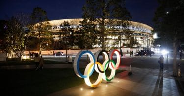 Uma estátua com os anéis olímpicos acesas durante a noite, e ao fundo um estádio iluminado em Tóquio.
