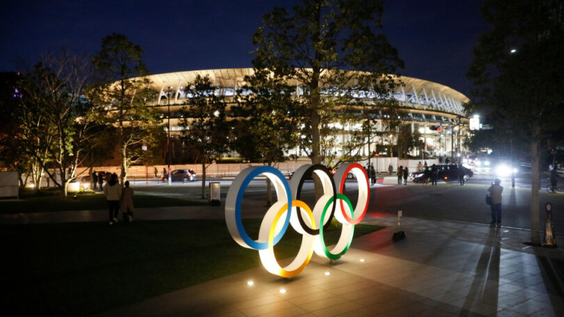 Uma estátua com os anéis olímpicos acesas durante a noite, e ao fundo um estádio iluminado em Tóquio.