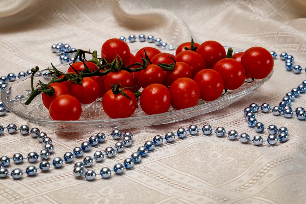Travessa de vidro cheia de tomates cereja. A travessa está sobre uma mesa forrada com uma linda toalha de cetim bege decorada com um colar de bolas prateadas.