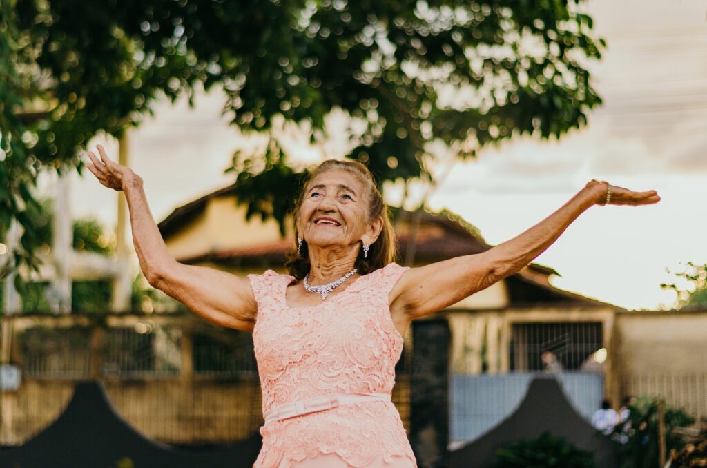 Mulher idosa de vestido sorrindo com as mãos para o alto, em uma rua, ao entardecer.