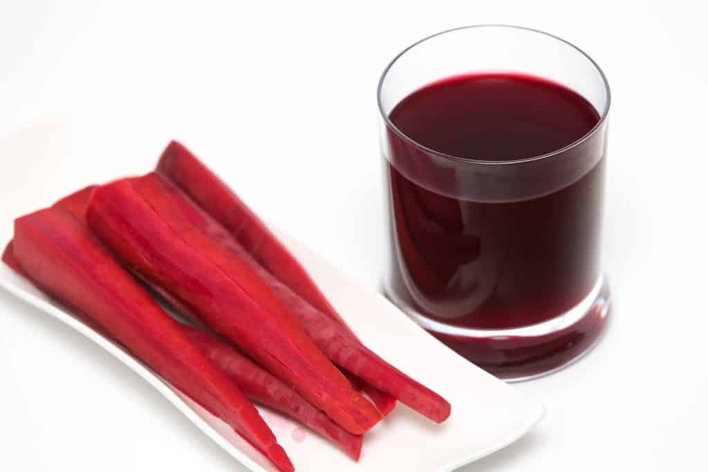 Imagem de uma bandeja com cenouras roxas já cortadas. Ao lado um copo pequeno de com suco da cenoura roxa.