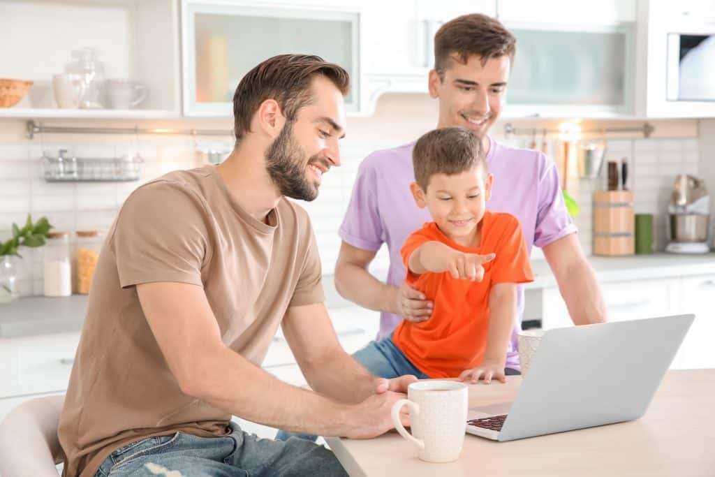 Dois homens de frente para um notebook em cima de uma bancada, com um menino pequeno sentado na bancada apontando para a tela.