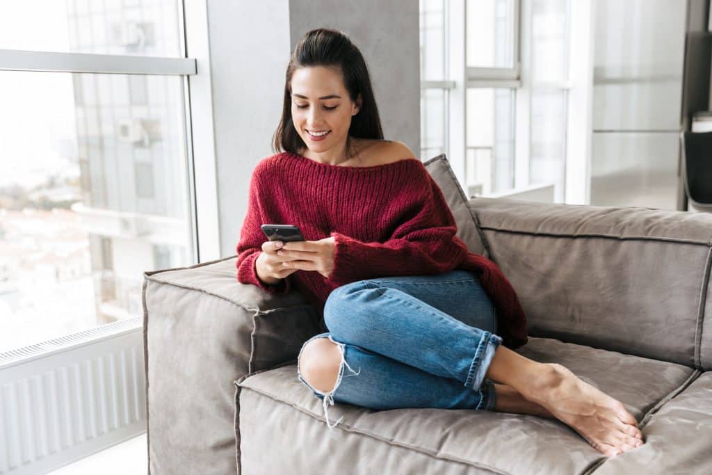 Mulher sentada no sofá segurando seu celular e sorrindo