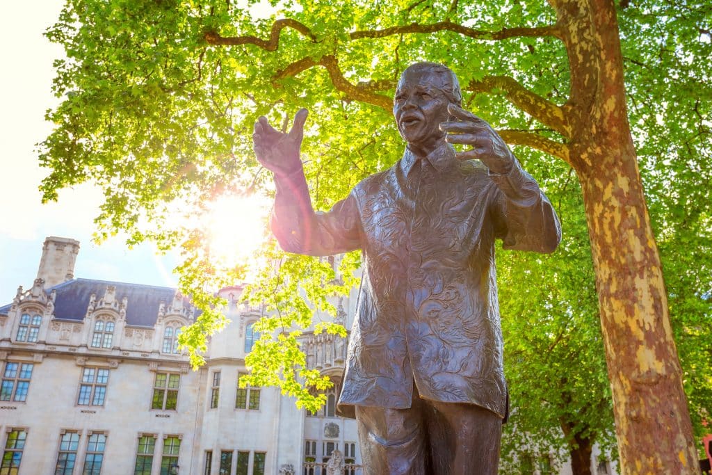 Imagem da estátua de Nelson Mandela sob a copa de uma linda árvore. Ao fundo um prédio antigo branco.