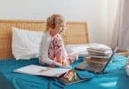 Menina sentada na cama com fones de ouvido olhando para um computador. Ao seu redor, tem-se livros, cadernos, canetas e uma calculadora.