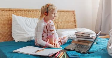 Menina sentada na cama com fones de ouvido olhando para um computador. Ao seu redor, tem-se livros, cadernos, canetas e uma calculadora.