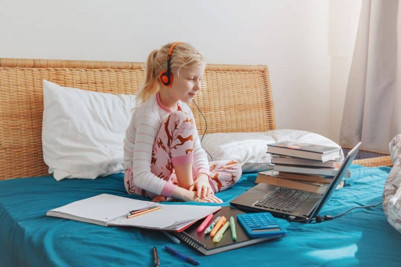 Menina sentada na cama com fones de ouvido olhando para um computador. Ao seu redor, tem-se livros, cadernos, canetas e uma calculadora.