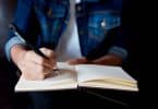 Imagem em detalhe de mulher escrevendo com lápis em um caderno. Ela usa camiseta e uma jaqueta jeans.