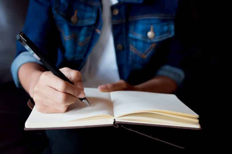 Imagem em detalhe de mulher escrevendo com lápis em um caderno. Ela usa camiseta e uma jaqueta jeans.