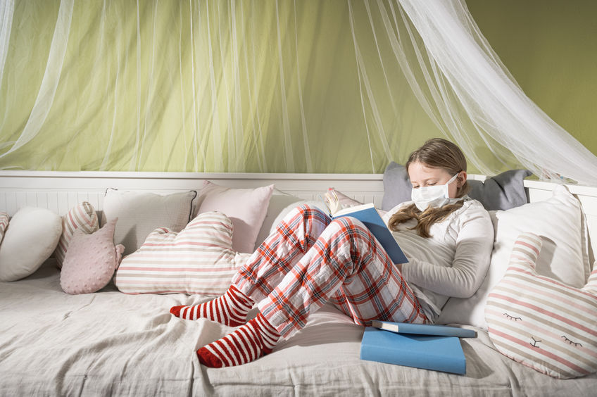 Menina usando máscara cirúrgica, sentada em cama com joelhos flexionados, lendo um livro.