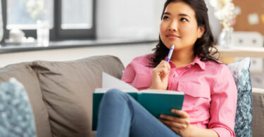 Mulher sentada em sofá e segurando caderno. Ela também segura uma caneta, que está encostada em seu queixo, com expressão pensativa.