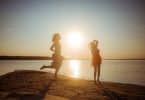 Mulher adulta e menina, ambas usando vestidos, pulando em praia durante o pôr do sol.