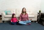 Mulher sentada no chão, encostada em sofá, meditando. Ao lado dela, uma menina pequena, imitando sua posição de meditação.