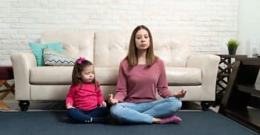 Mulher sentada no chão, encostada em sofá, meditando. Ao lado dela, uma menina pequena, imitando sua posição de meditação.