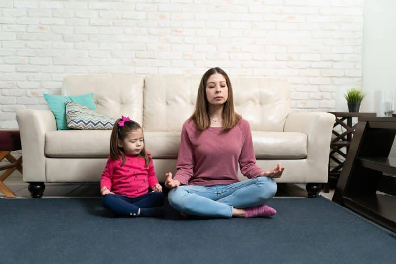 Mulher sentada no chão, encostada em sofá, meditando. Ao lado dela, uma menina pequena, imitando sua posição de meditação.