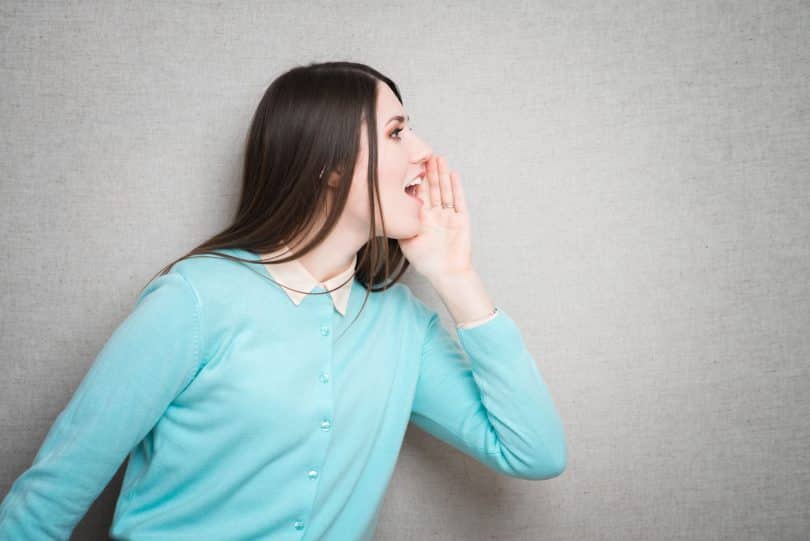 Mulher de perfil em frente a parede cinza, com uma mão encostada na parte do rosto mais distante da câmera, e com a boca aberta, como se estivesse gritando.