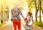 Menina pequena com cabelo preso e usando uma mochila nas costas, de mãos dadas com uma mulher. Elas estão andando, e de costas para a câmera.