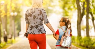 Menina pequena com cabelo preso e usando uma mochila nas costas, de mãos dadas com uma mulher. Elas estão andando, e de costas para a câmera.