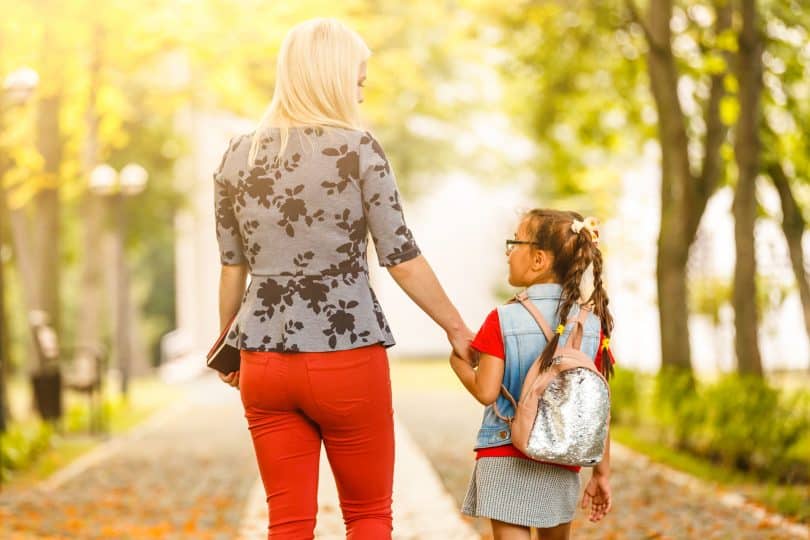 Menina pequena com cabelo preso e usando uma mochila nas costas, de mãos dadas com uma mulher. Elas estão andando, e de costas para a câmera.