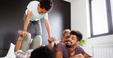 Família feliz em uma cama. Uma mulher está deitada de costas, com as pernas flexionadas, segurando as mãos e equilibrando um menino em suas pernas. Ao lado, uma menina deitada nas costas de um homem olham para a situação e dão risada.