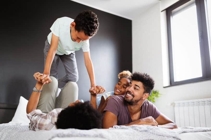 Família feliz em uma cama. Uma mulher está deitada de costas, com as pernas flexionadas, segurando as mãos e equilibrando um menino em suas pernas. Ao lado, uma menina deitada nas costas de um homem olham para a situação e dão risada.