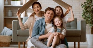 Família reunida em sala, segurando papelão sobre suas cabeças, simbolizando um telhado. Uma mulher e uma menina nas pontas seguram o papelão, com um homem e uma menina em seu colo no centro.