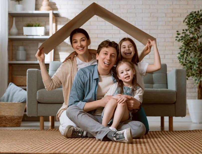 Família reunida em sala, segurando papelão sobre suas cabeças, simbolizando um telhado. Uma mulher e uma menina nas pontas seguram o papelão, com um homem e uma menina em seu colo no centro.
