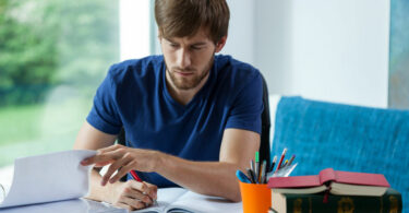 Homem sentando em frente de um caderno, escrevendo nele.