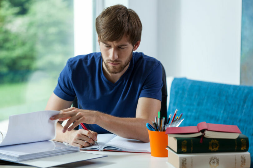 Homem sentando em frente de um caderno, escrevendo nele.