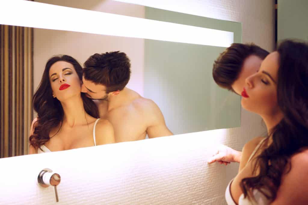 Imagem de um casal de homem e mulher. Eles estão dentro de um quarto de frente para um espelho. Ele beija o pescoço da sua companheira.