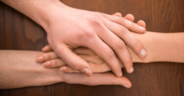 Duas mãos de uma pessoa segurando a mão de outra, sobre uma mesa de madeira