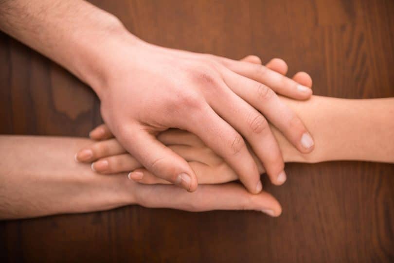 Duas mãos de uma pessoa segurando a mão de outra, sobre uma mesa de madeira
