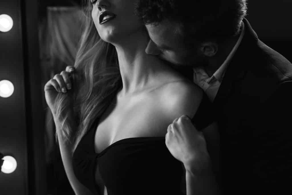 Imagem de um casal (homem e mulher). Ele veste um vestido sexy preto. Ele beija o pescoço dela de forma carinhosa.