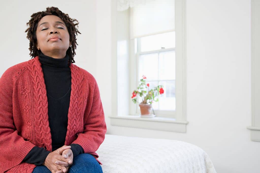 Mulher sentada na cama, em um quarto, com os olhos fechados e as mãos cruzadas sobre o colo.