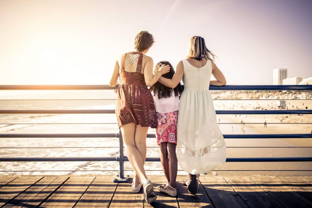 Duas mulheres de pé, com uma menina entre elas. Elas estão em um pier, de costas para a câmera, observando o mar. As duas mulheres abraçam a menina.