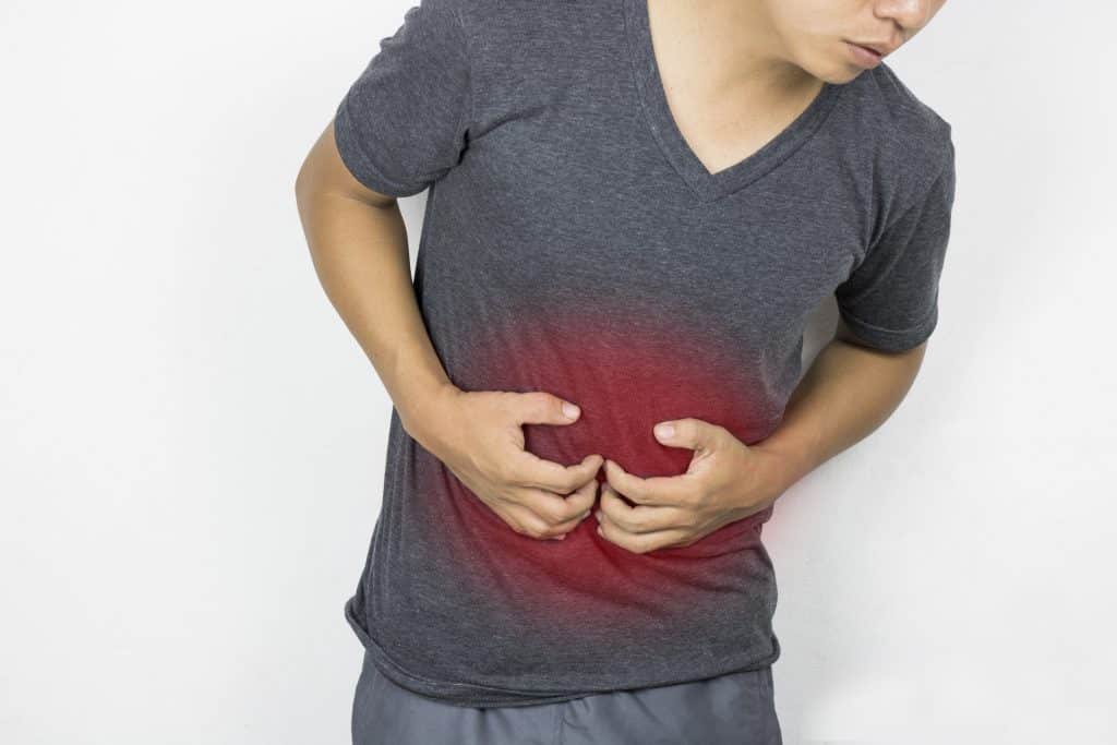 Imagem de um homem jovem vestindo uma camiseta cinza escura. Ele está com dores do fígado e suas mãos estão sobre ele.
