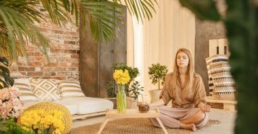 Mulher sentada no chão, de olhos fechados, meditando em sua sala e estar.