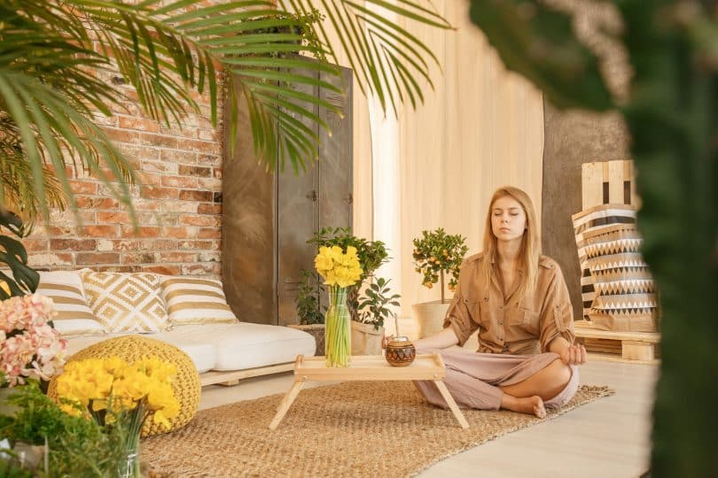 Mulher sentada no chão, de olhos fechados, meditando em sua sala e estar.