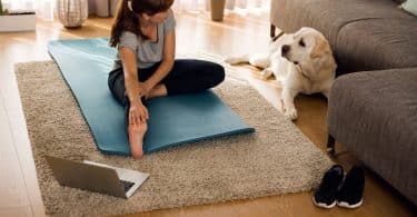 Mulher sentada no chão da sala de estar, alongando-se, ao lado de seu cachorro e de um laptop.