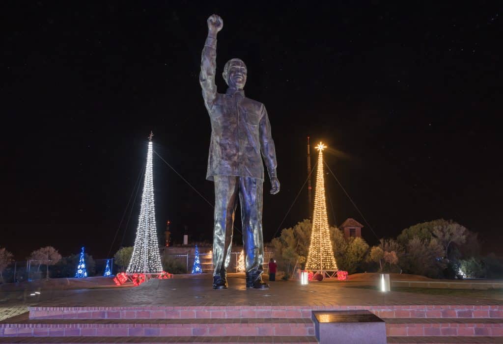 Imagem de uma estátua gigante de Nelson Mandela instalada em uma das praças da África. Ao lado da estátua duas árvores de Natal iluminadas.