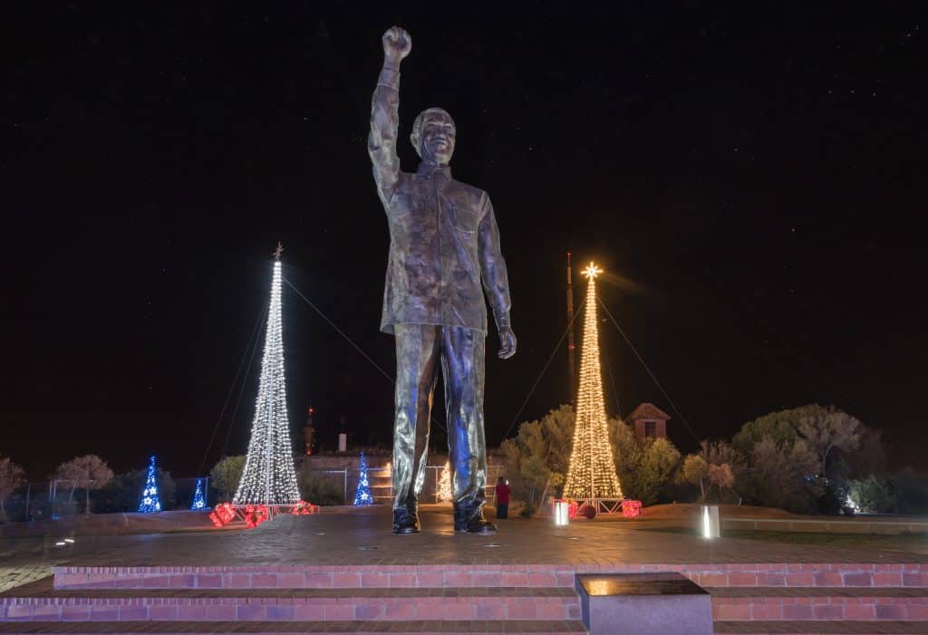 Imagem de uma estátua gigante de Nelson Mandela instalada em uma das praças da África. Ao lado da estátua duas árvores de Natal iluminadas.
