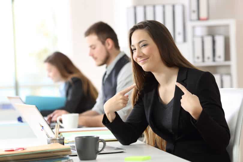 Mulher no trabalho sorrindo e apontando para si mesma