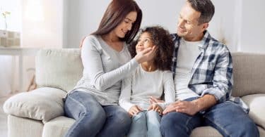 Homem e mulher sentados em sofá. Entre eles, uma menina está sentada. A mulher tem sua mão direita no rosto da menina e elas se olham. O homem observa as duas. Todos sorriem.