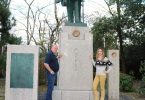 Um homem e uma mulher ao lado de uma estátua de um homem, com pedestal alto.