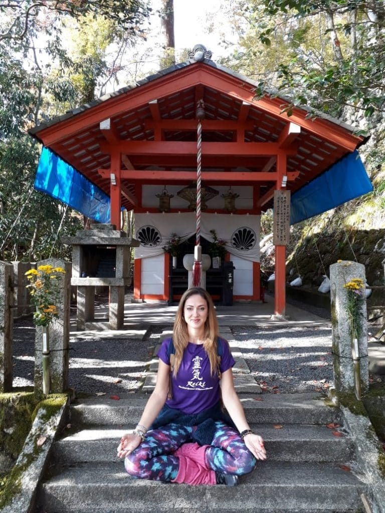 Mulher sentada com as pernas cruzadas em escada, em frente a um pequeno templo.