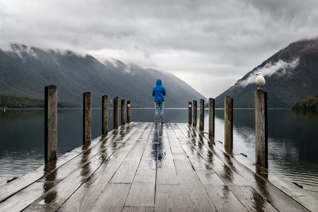 Homem em pé em deck de madeira de costas com montanhas ao fundo