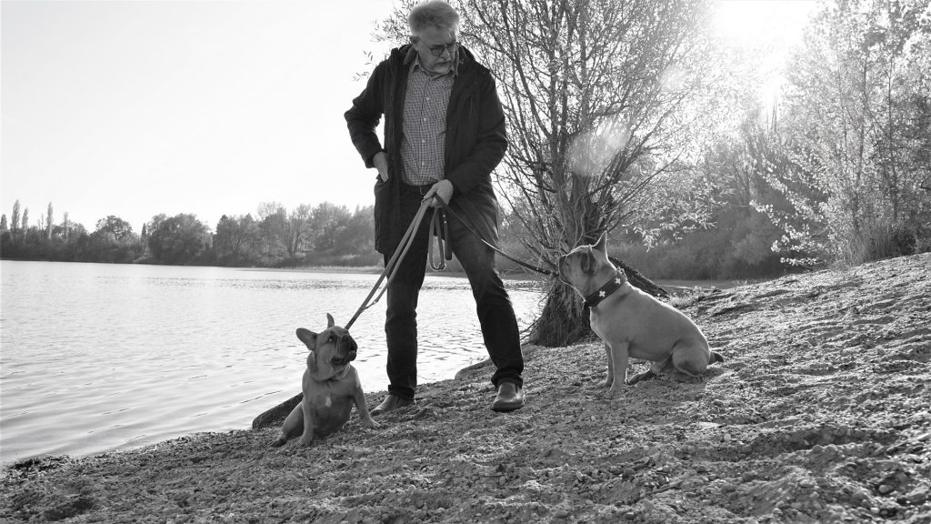 Imagem em preto e branco de um homem em um lago segurando em suas duas guias com os seus cachorros de estimação.
